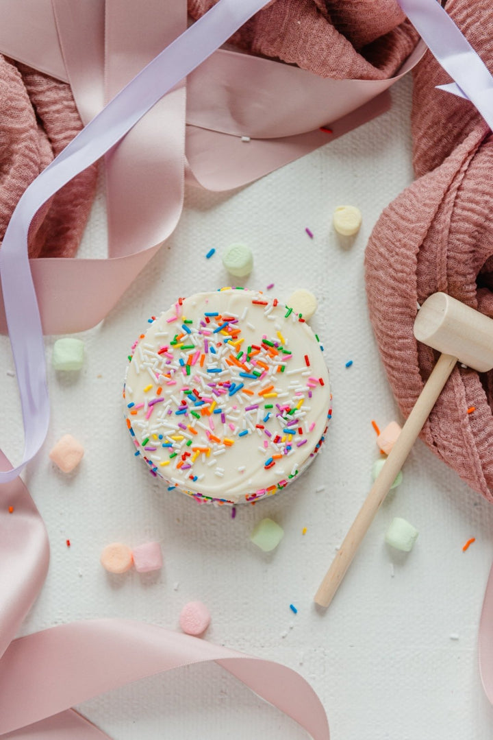Decorative cookie with colorful sprinkles on a white surface with pink ribbons and a wooden mallet.