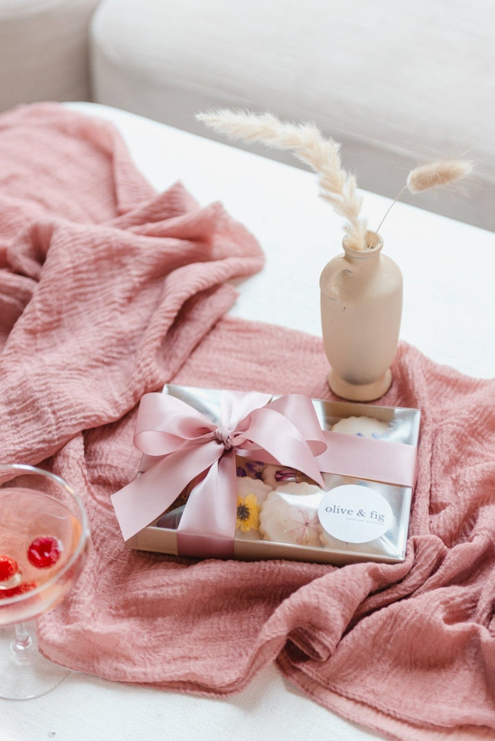 Pink blanket draped over a surface with a glass of red wine, a book, and a vase with dried flowers.