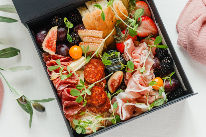 Assorted meats, cheeses, fruits, and vegetables on a black platter with a pink knitted item in the background.