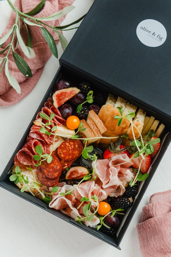 Gourmet charcuterie board with meats, cheeses, and fruits in a black box labeled 'Olive & Fig'.