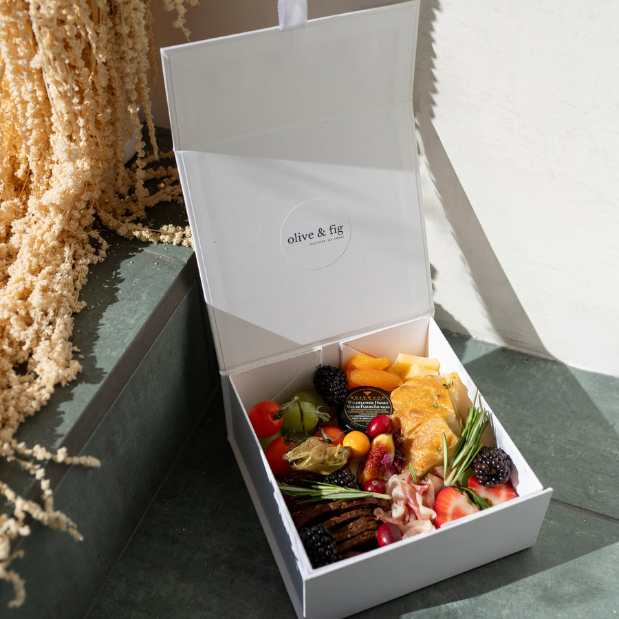 Open white charcuterie gift box with assorted treats on a textured surface