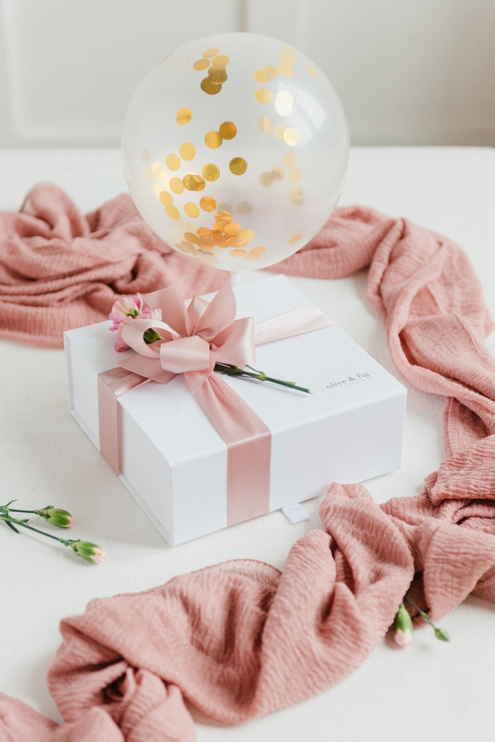 White gift box with pink ribbon and flower, surrounded by pink fabric and a clear balloon with gold confetti on a white surface.