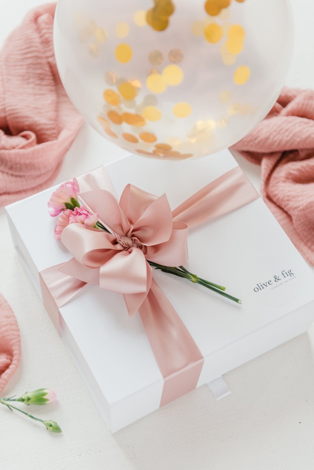 Gift box with pink ribbon and flowers, surrounded by pink fabric and a balloon with gold confetti on a light background.