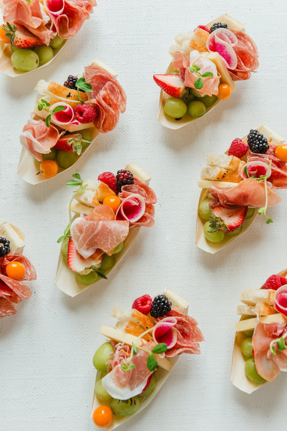 Small appetizers with prosciutto, fruits, and vegetables on a white surface