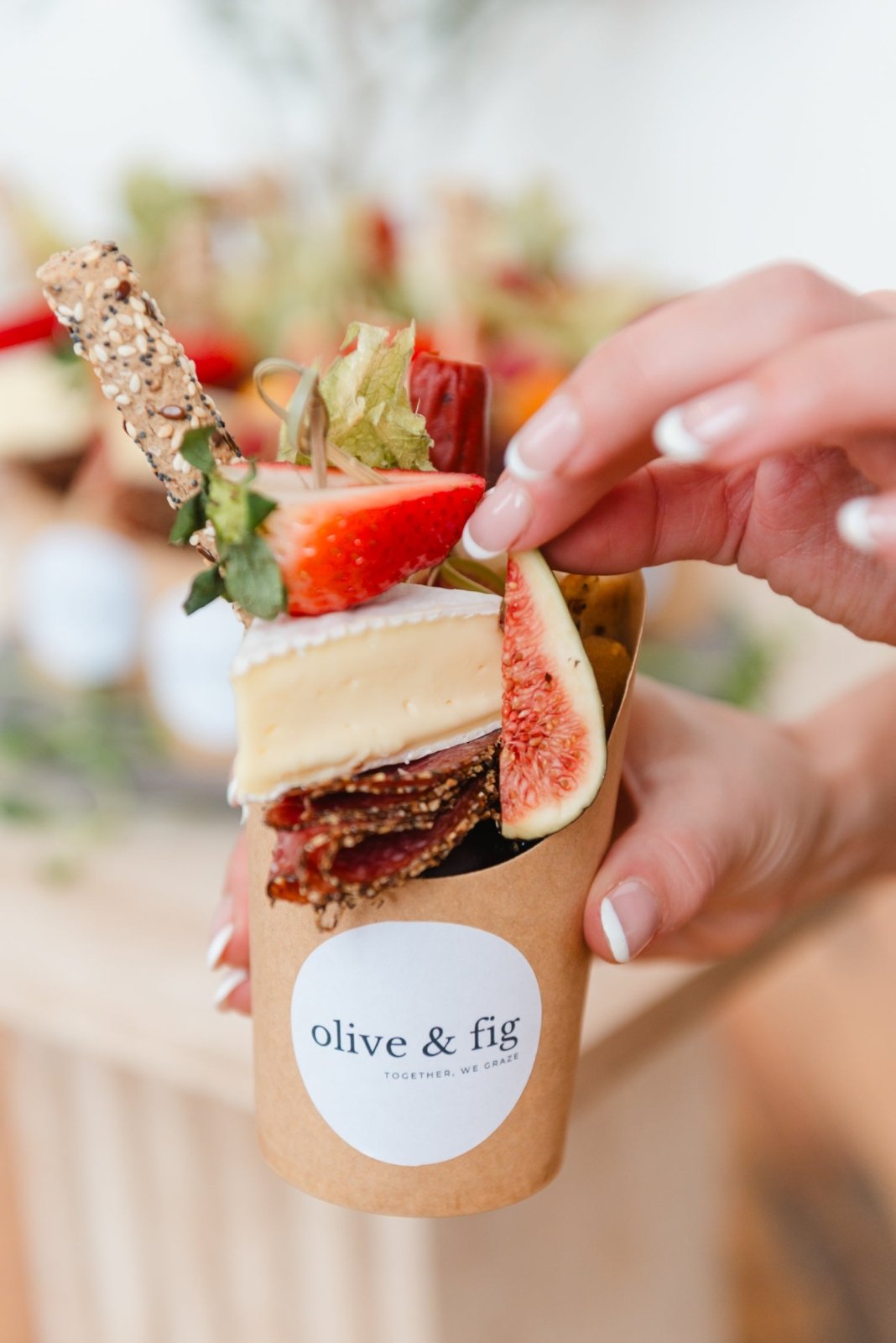 Hand holding a paper cup with a snack and 'olive & fig' label, blurred background