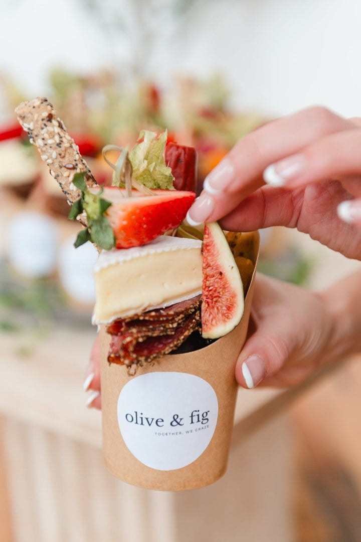Hand holding a paper cup with a snack and 'olive & fig' label, blurred background
