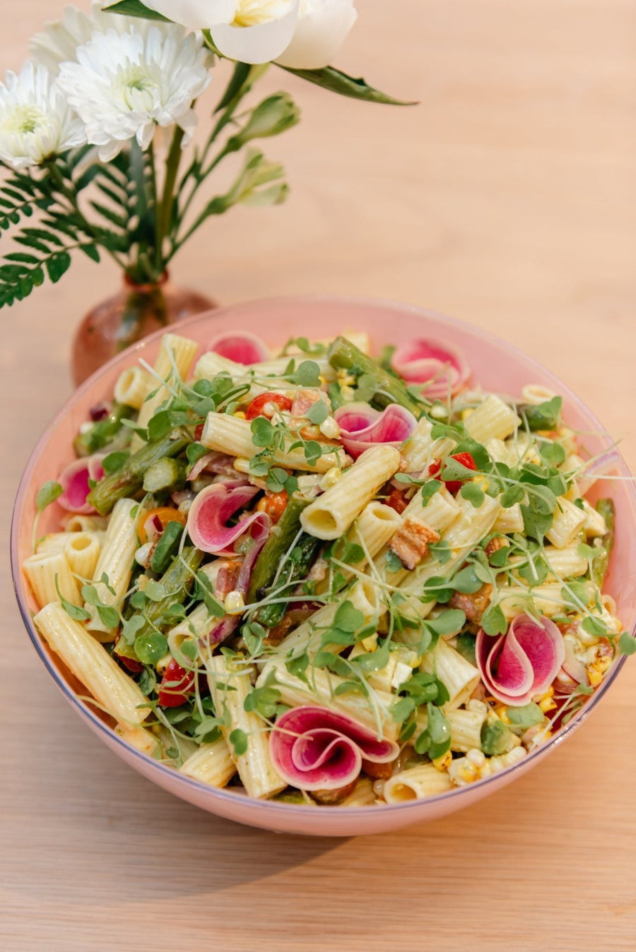 Pasta salad with vegetables in a pink bowl on a wooden table, accompanied by white flowers.