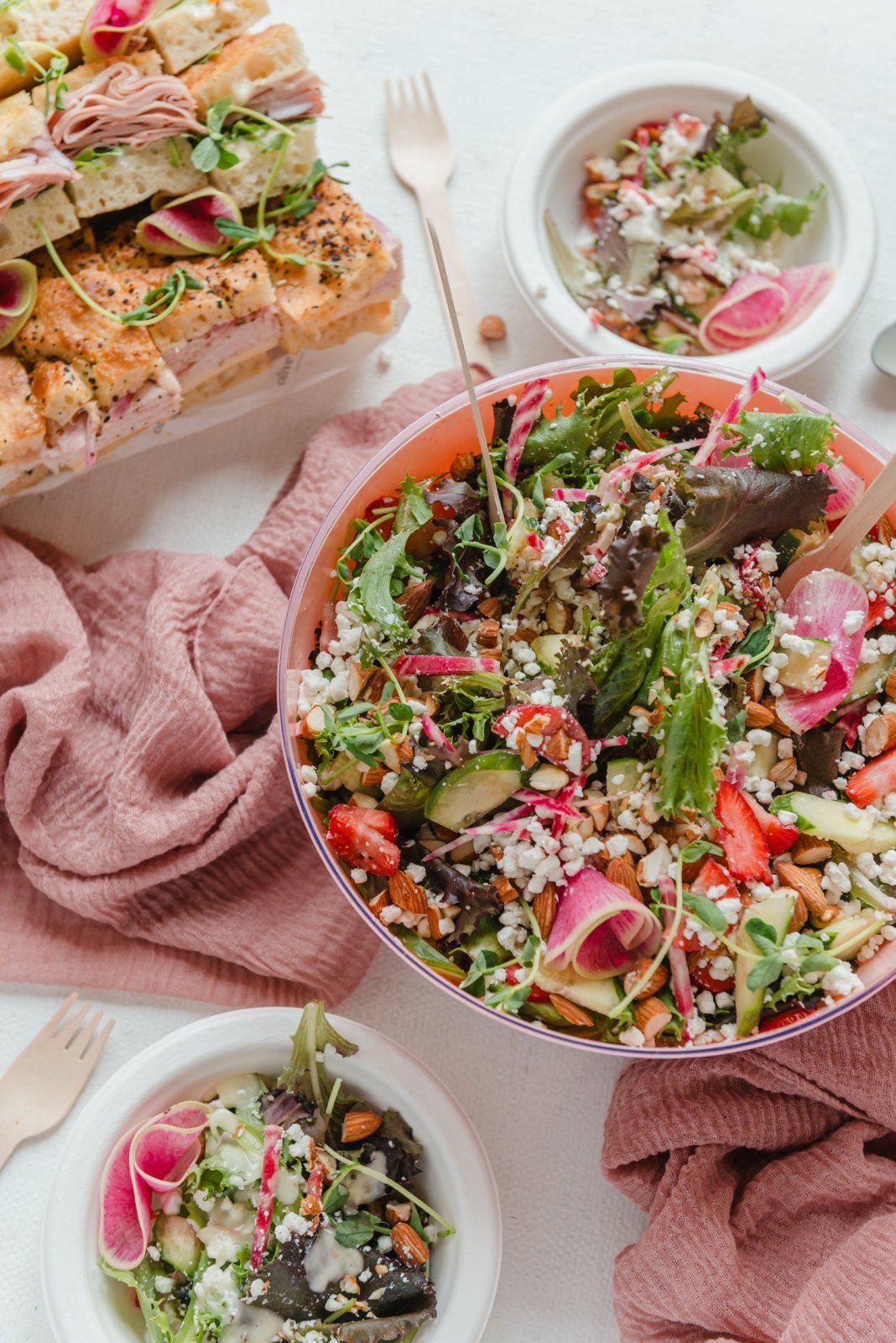 Colorful salad in a bowl with a pink napkin and sandwich in the background on a white surface.