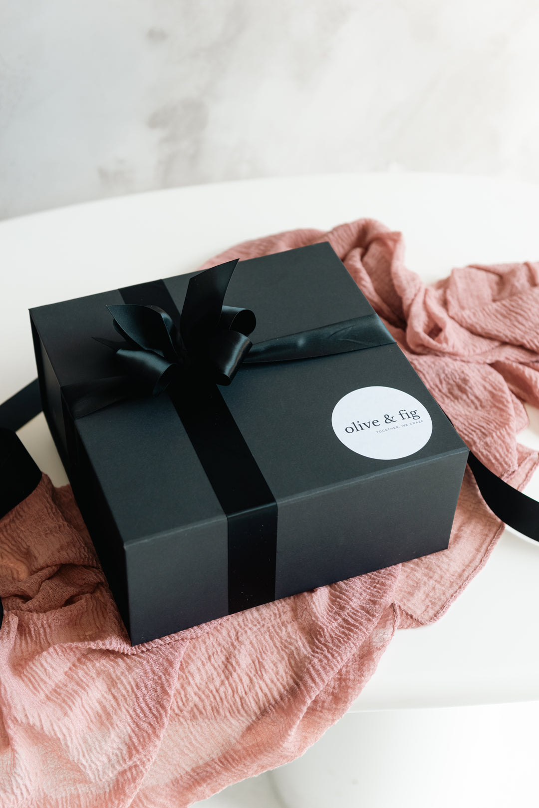 Black gift box with a bow and 'olive & fig' label on a pink fabric background