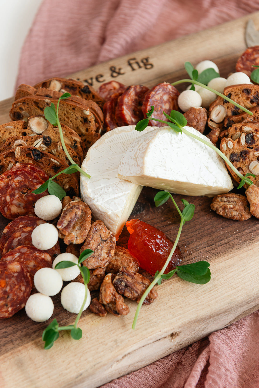 Wooden board with assorted meats, cheeses, and bread on a pink cloth.