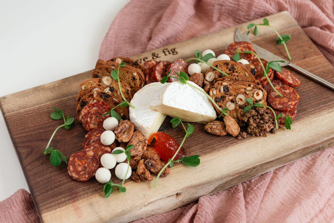 Wooden charcuterie board with cured meats, cheeses, and nuts on a pink cloth.