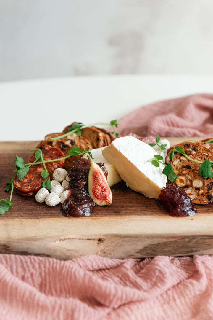 Wooden board with assorted fruits, nuts, and cheese on a pink cloth.