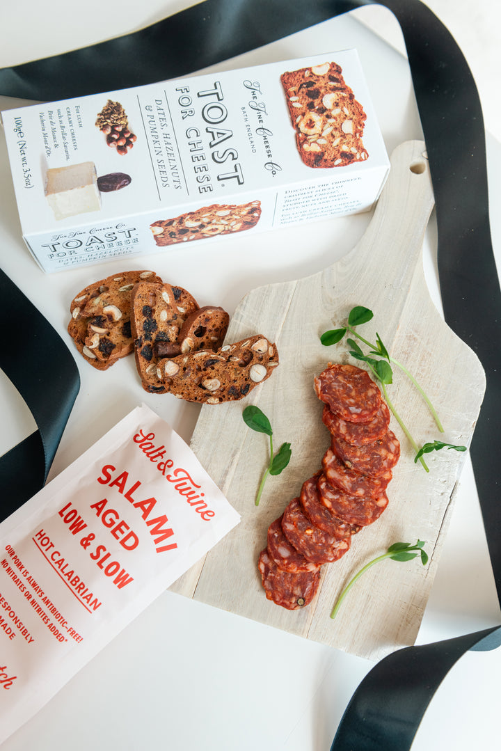 Packaged salami, crackers, and a box of Toast & Cheese on a white surface with black handles.