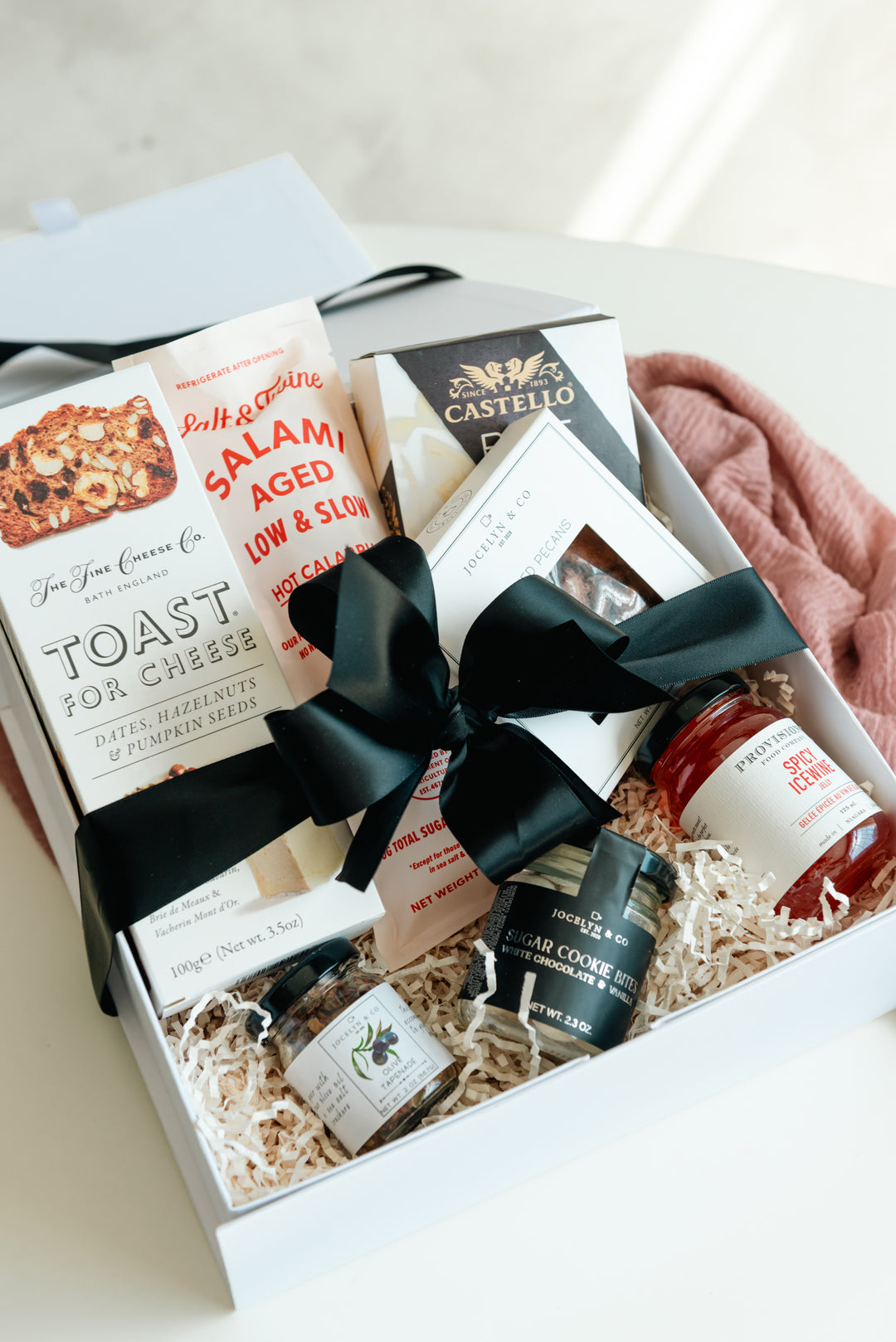 Gift box with assorted food items and a black bow on a light background