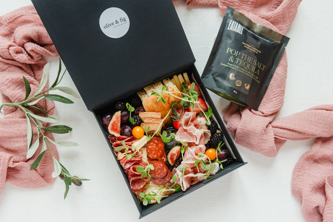 Gourmet food box with a variety of snacks and a small bag on a white surface with pink fabric.